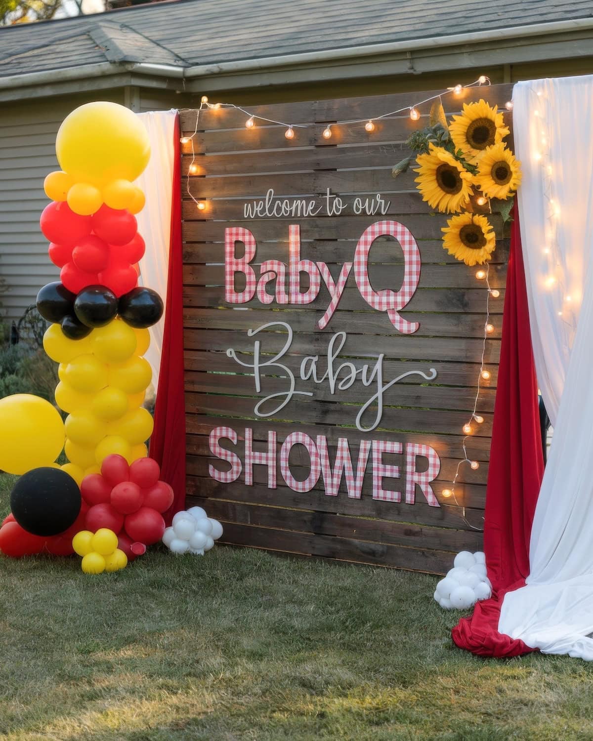 A wooden backdrop on a lawn displays the phrase "Welcome to our BBQ BabyQ Baby Shower" in pink and white letters. Surrounding it are red, black, and yellow balloon clusters, sunflower decorations, and white and red drapes with string lights.