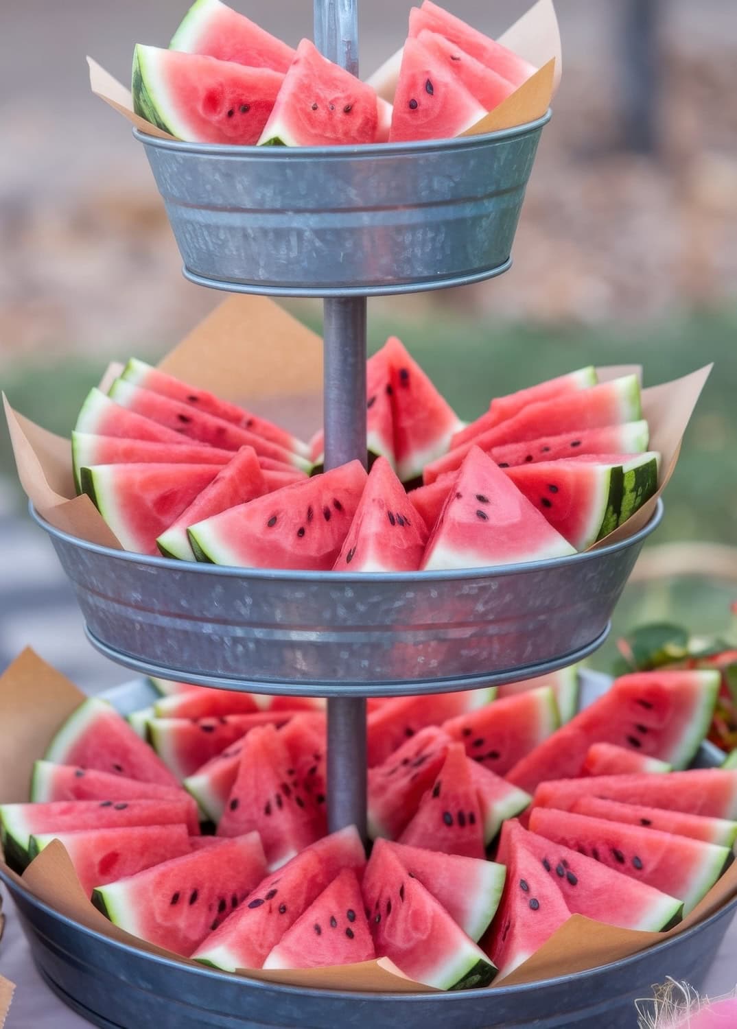 A three-tiered metal stand elegantly showcases triangular watermelon slices at this BBQ-themed baby shower. Each tier brims with vibrant red and green pieces on brown paper, adorned with black seeds. The softly blurred background adds a delightful touch to the scene.