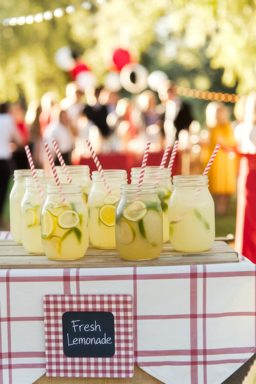 Mason jars filled with lemonade and lemon slices, each with a red and white striped straw, grace a table adorned with a checkered cloth at the BBQ baby shower. A small chalkboard sign reads "Fresh Lemonade." Blurred background shows people and festive decorations.