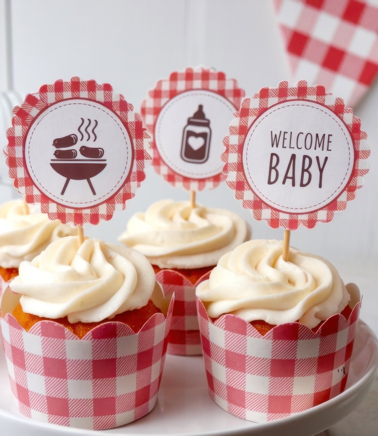 Four cupcakes with swirl frosting sit on a white plate. Each has a red and white gingham wrapper and a toothpick topper. The toppers, for a BBQ baby shower, read "WELCOME BABY" and feature images of a grill and baby bottle.