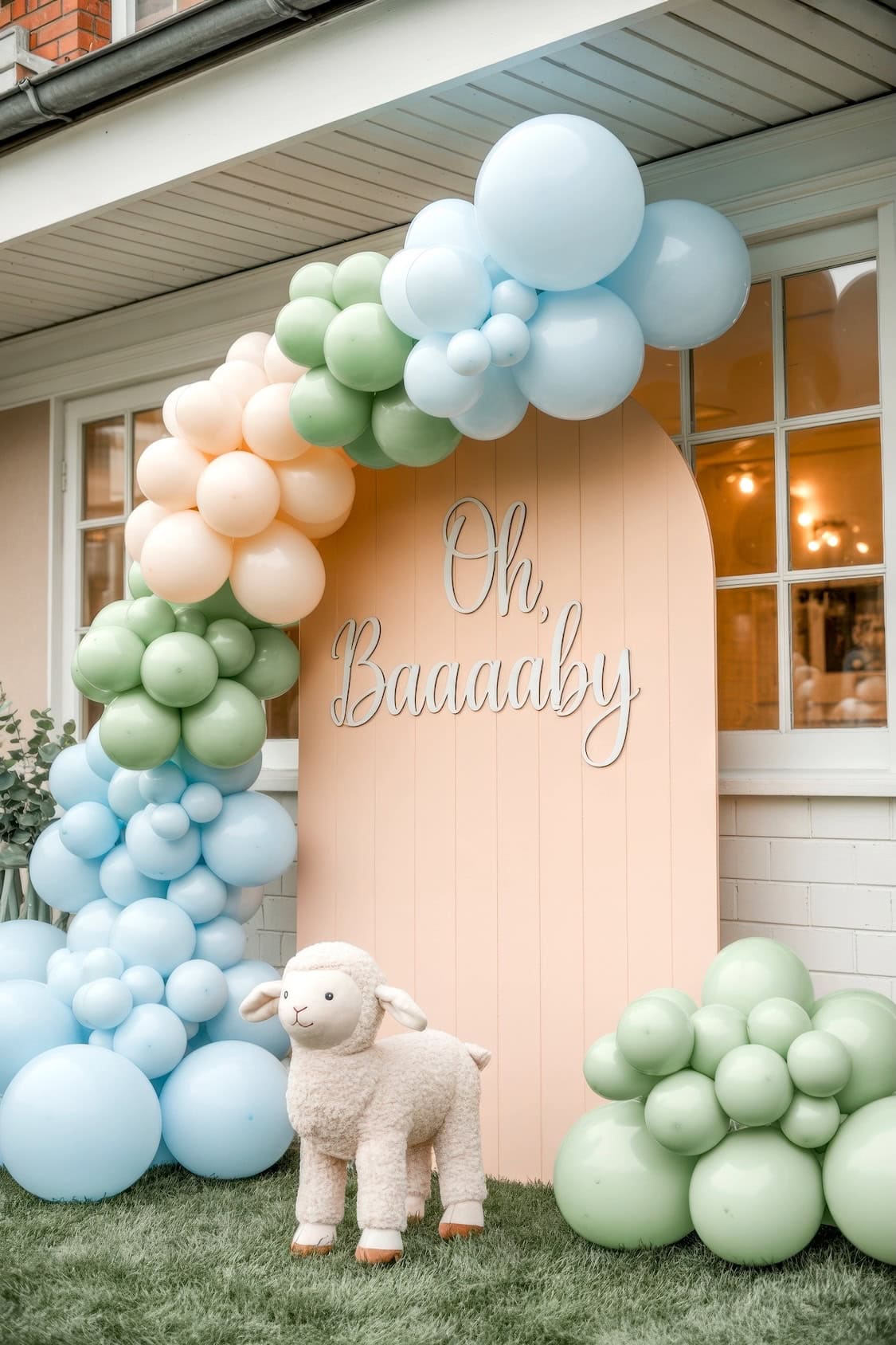 A pastel balloon arch in blue, green, and peach frames a peach-colored panel with “Oh, Baaaaby” written on it. Perfect for summer baby shower ideas, a plush lamb toy stands in front, all set outdoors on grass near a house.