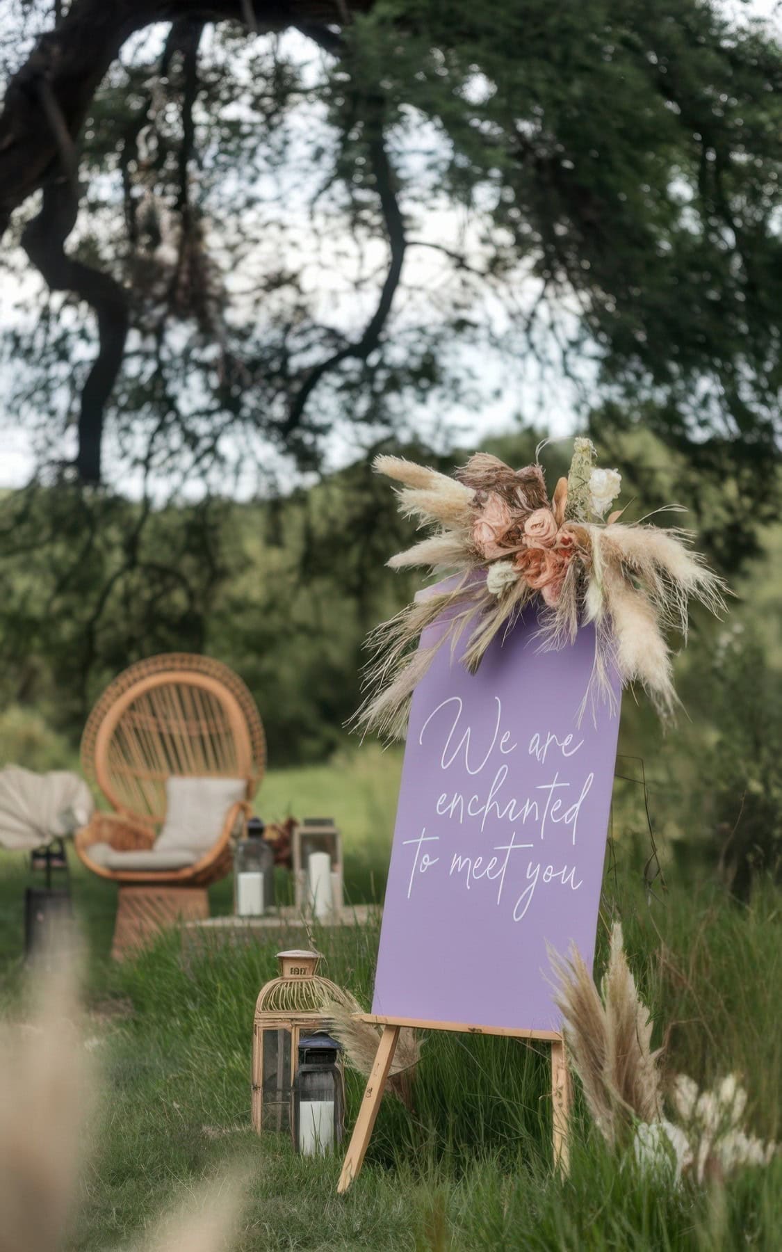 A decorative lavender sign with the words "We are enchanted to meet you" stands on an easel in a grassy outdoor setting—perfect for summer baby shower ideas—adorned with pampas grass, flowers, a wicker chair, and lanterns in the background.