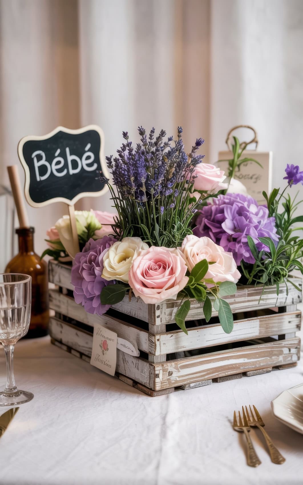 A rustic wooden crate filled with pink roses, lavender, and purple flowers sits on a table with wine glasses and cutlery—a charming touch for summer baby shower ideas. A small chalkboard sign reading "Bébé" nestles among the blooms.