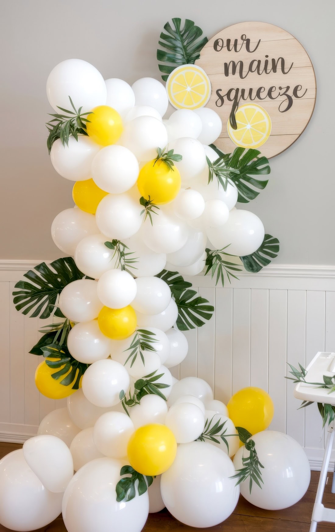 A decorative arrangement of white and yellow balloons with green leaves stands next to a wooden sign that reads "our main squeeze" with lemon slice illustrations—perfect for summer baby shower ideas—against a white wall.