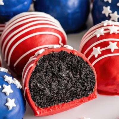 Red, white, and blue cake balls decorated with stripes and stars; one is cut in half to reveal a dark chocolate interior. These Red White and Blue Oreo Balls evoke an American patriotic theme.