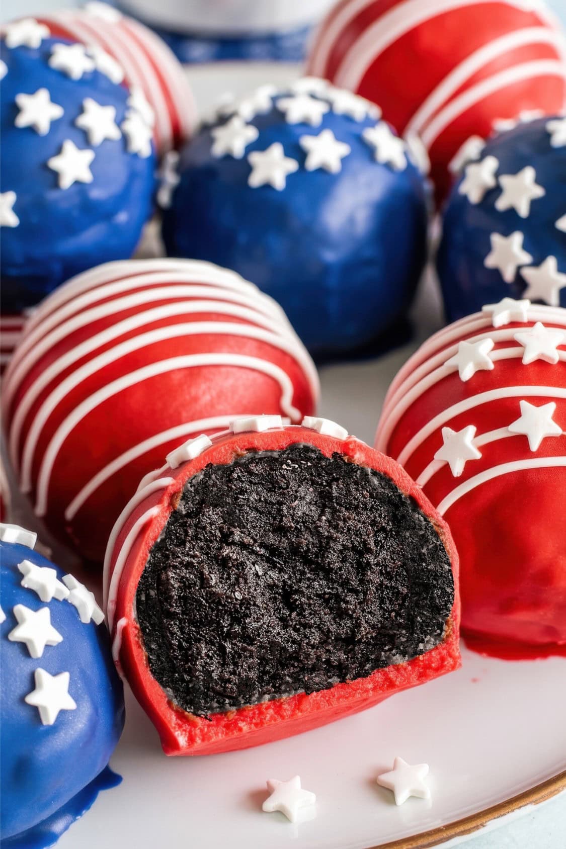 A plate of Red White and Blue Oreo Balls coated in vibrant red and blue icing, decorated with white stars and stripes. One red Oreo ball is cut in half, revealing a rich dark chocolate interior.