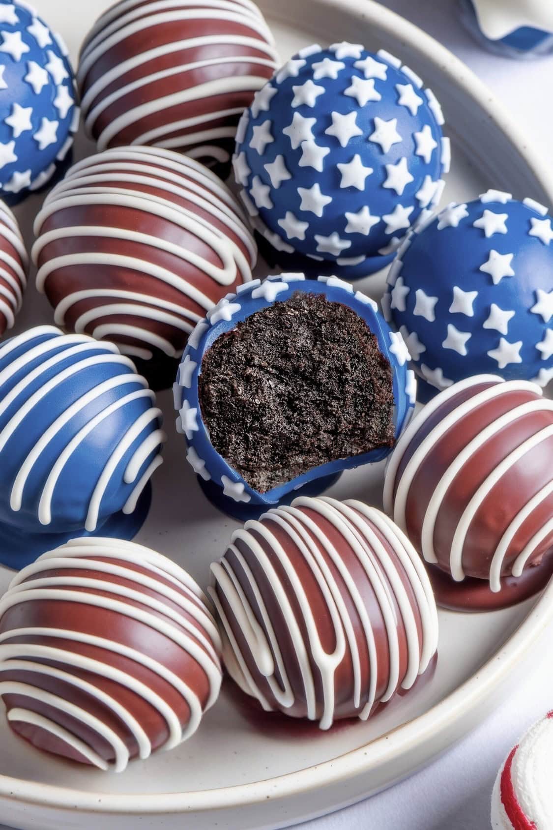 A plate of round Red White and Blue Oreo Balls is decorated with red, white, and blue icing; some feature white star patterns while others have stripes. One ball has a bite taken out, revealing a rich dark chocolate center.