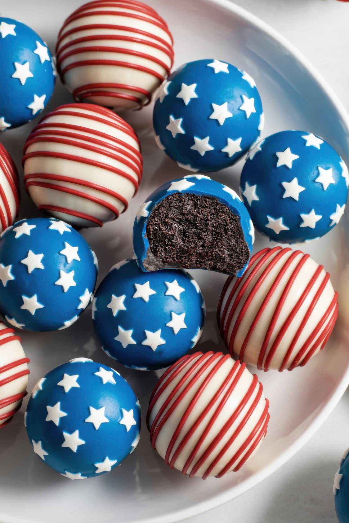 A plate of Red White and Blue Oreo Balls decorated with blue icing and white stars, plus red and white stripes. One Oreo ball has a bite taken out, revealing a chocolate interior. The colors evoke a patriotic, American flag theme.