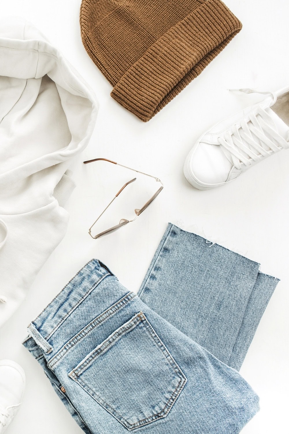 Flat lay of casual clothing and accessories, curated in a Stitch Fix style: a brown knit beanie, cream hoodie, white sneakers, blue jeans, and sunglasses arranged neatly on a white background.