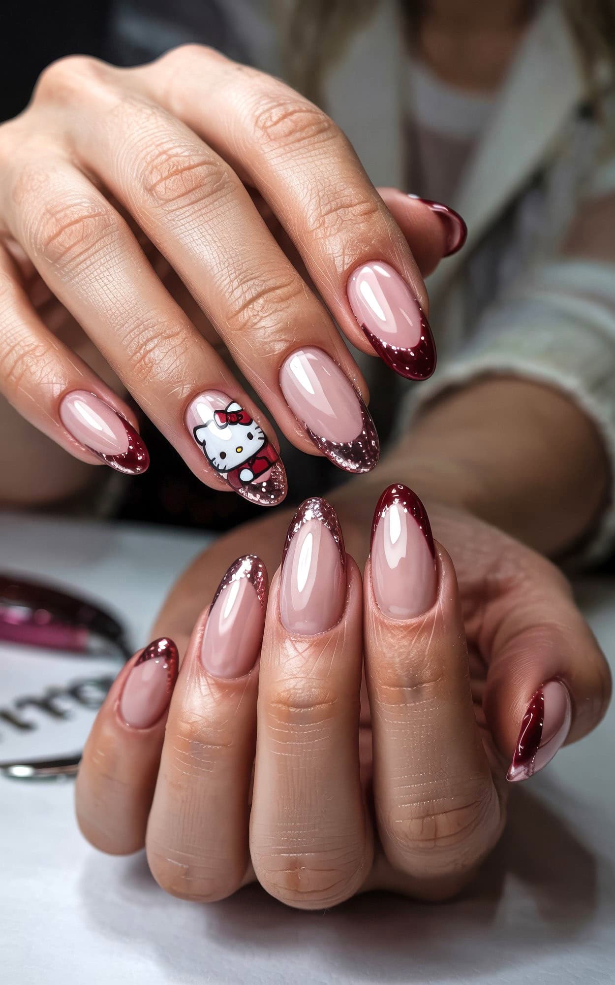 A close-up of hands with almond-shaped nails featuring a pink base, dark red glittery French tips, and one accent nail with a festive Hello Kitty Christmas nails design.