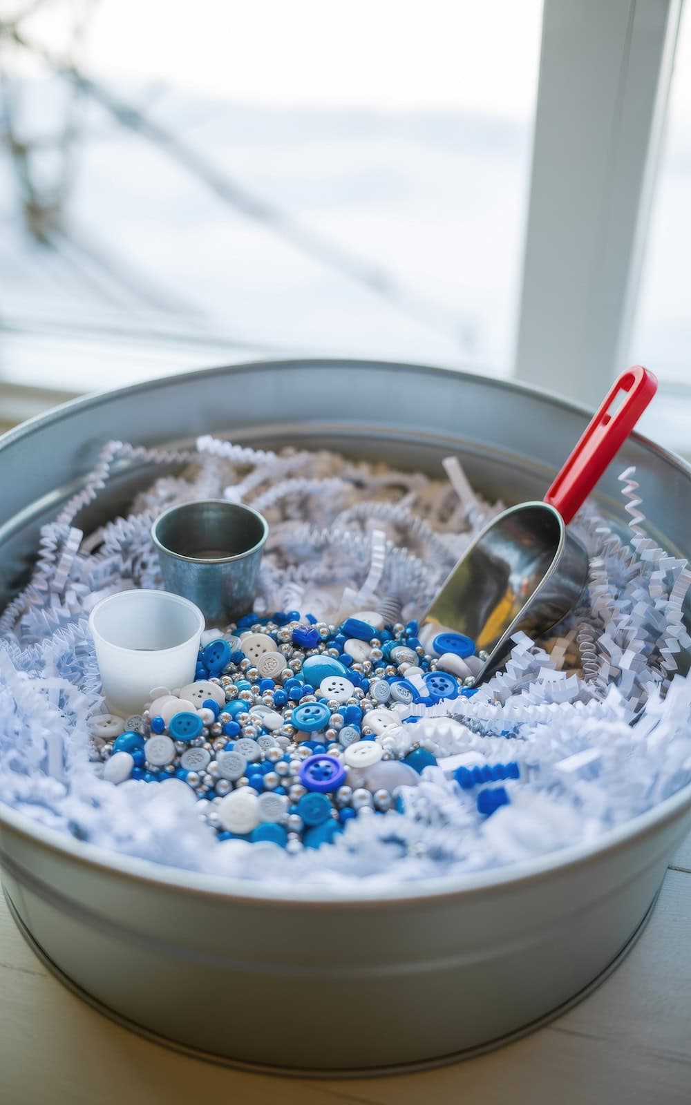 A round metal tray filled with white paper shreds, blue and white buttons, small beads, and a metal scoop with a red handle creates inviting winter sensory bins, accompanied by a small metal cup and plastic cup, all placed near a window.