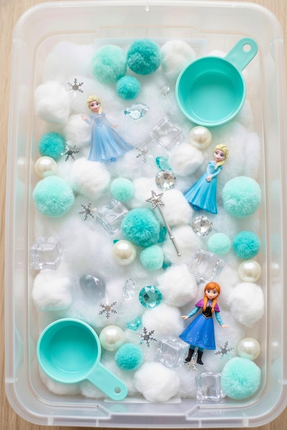 A winter sensory bin filled with white and blue pom-poms, toy snowflakes, plastic gems, pearls, miniature Elsa and Anna dolls, and teal scoops, all arranged in a clear container on a light surface.