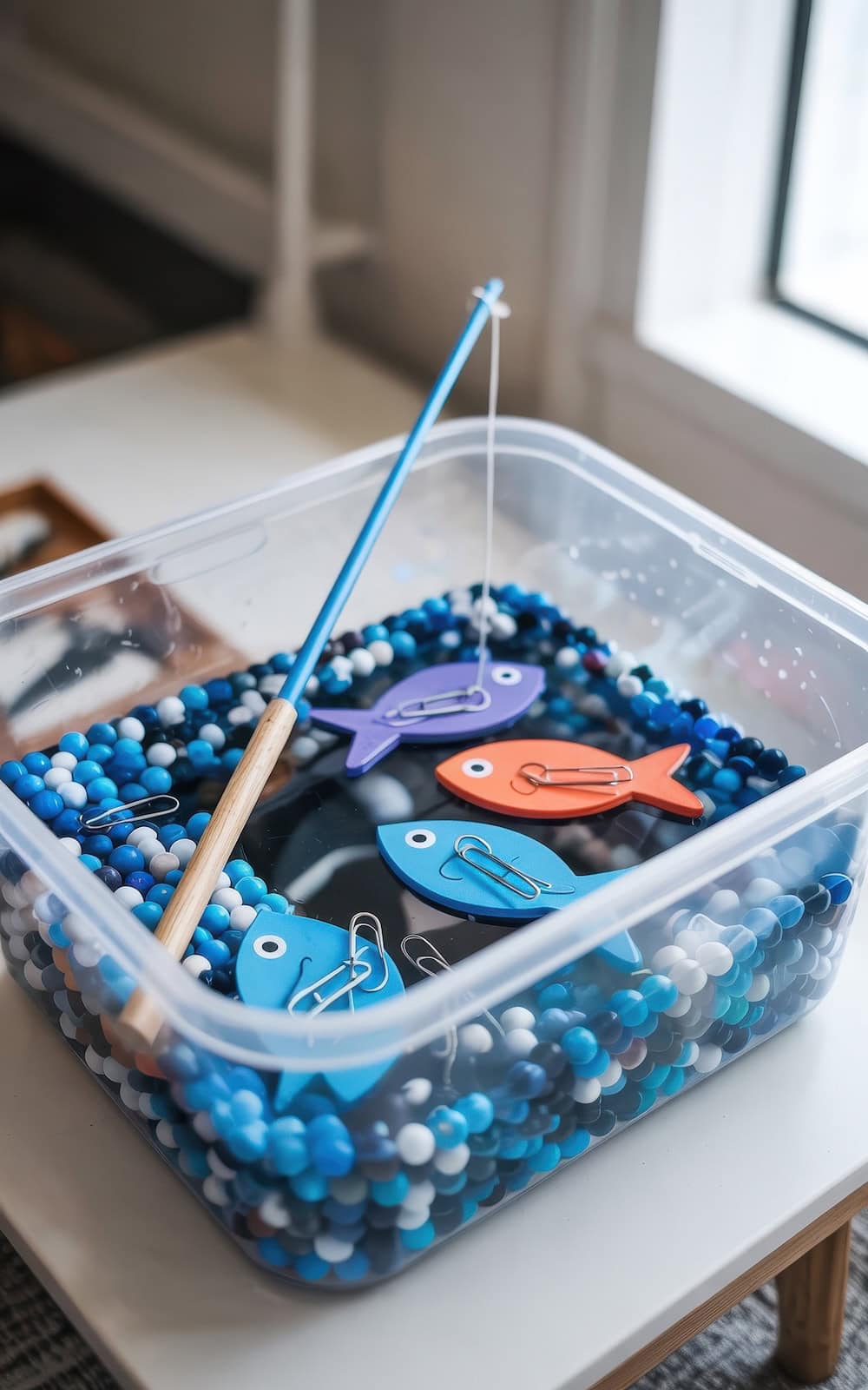 A plastic bin filled with blue, black, and white beads holds colorful wooden toy fish and a fishing rod, creating a winter sensory bin that simulates a fun fishing game for children.