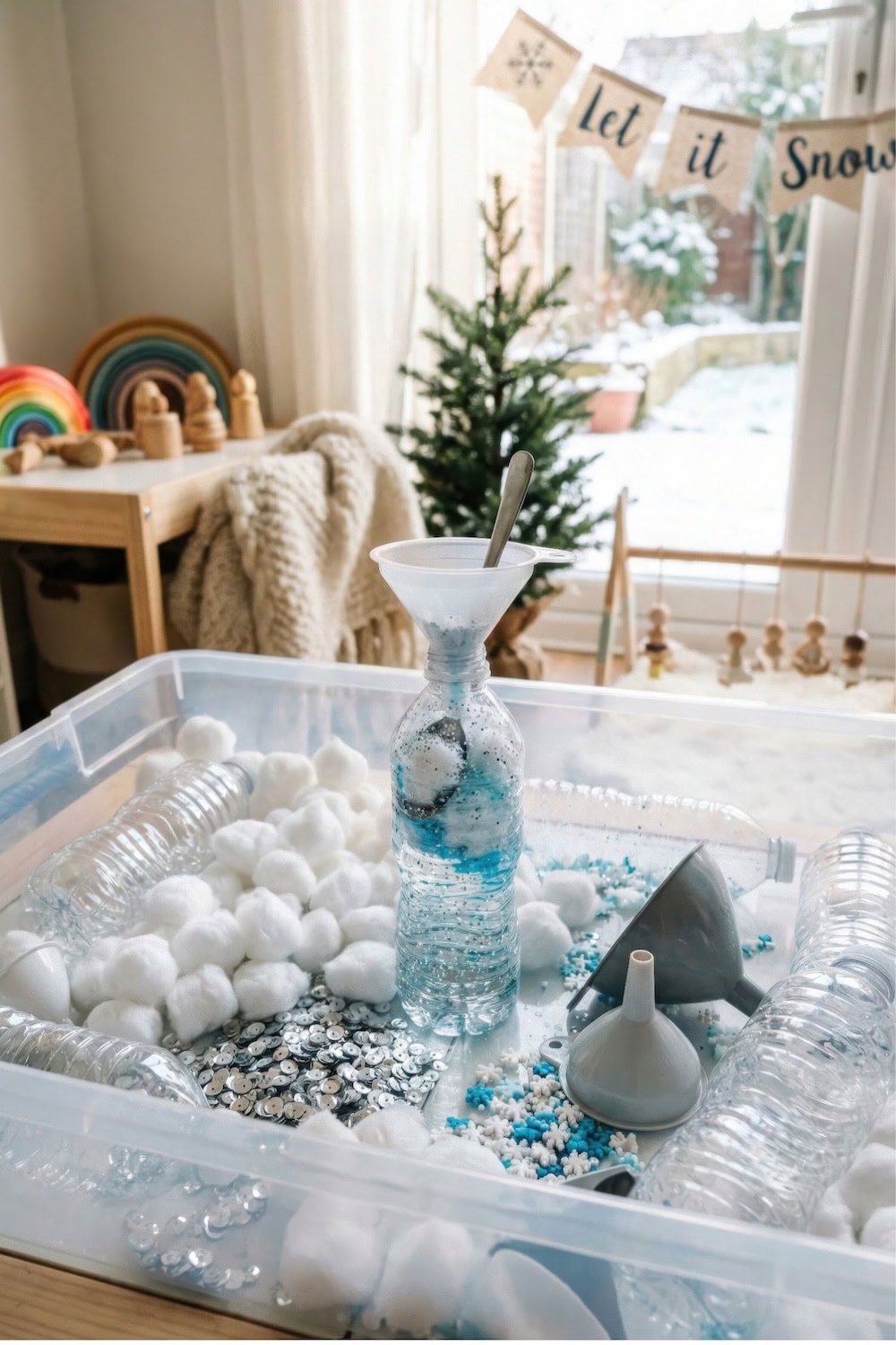A winter sensory bin filled with cotton balls, blue rice, silver coins, funnels, and a plastic bottle sits on a table near a window. A small Christmas tree and "Let it Snow" banner add festive charm in the background.
