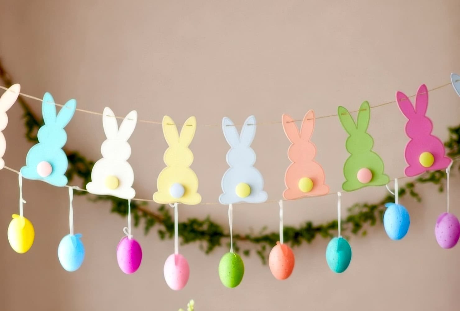 A festive garland of pastel-colored paper bunnies with cotton tails, perfect for a bunny baby shower, hangs above colorful plastic Easter eggs, strung across a blurred background with greenery.
