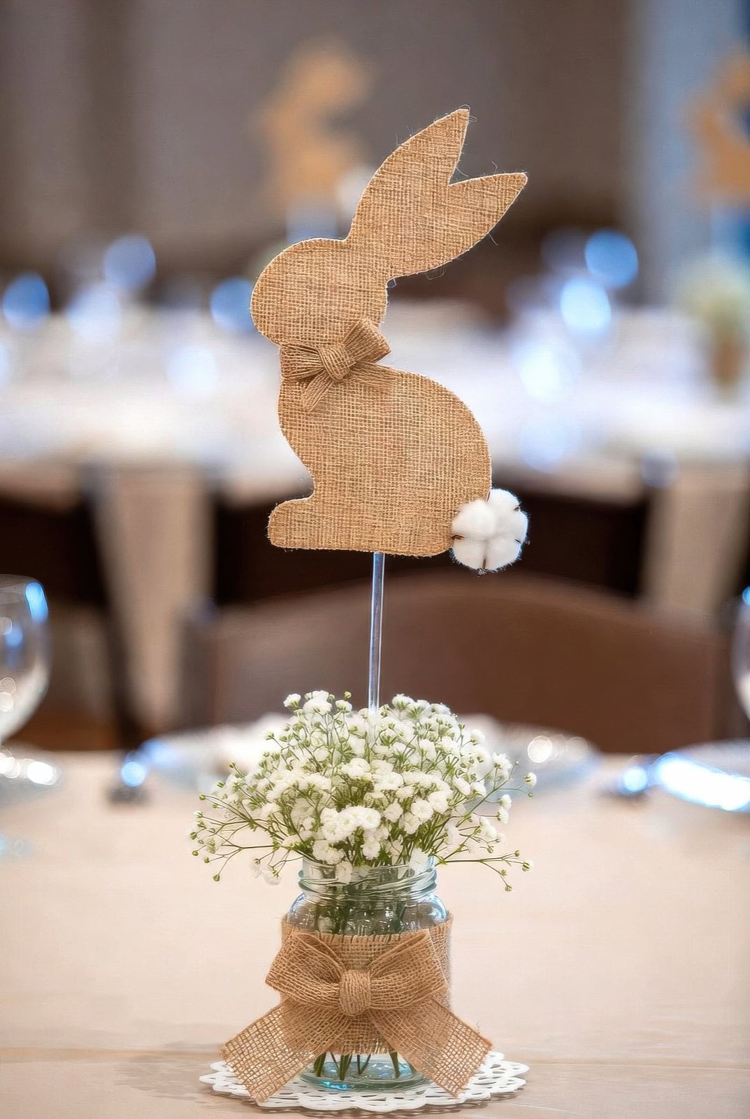 A burlap bunny silhouette with a bow and pom-pom tail on a stick is arranged in a jar of white flowers, tied with a burlap bow, serving as a charming Bunny baby shower table centerpiece.