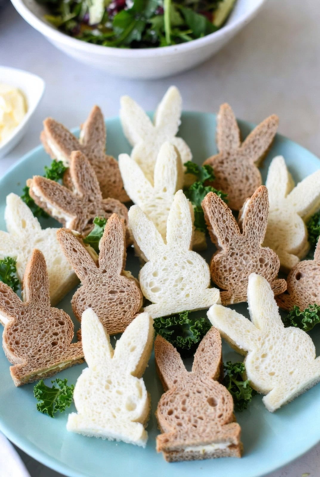 A plate of bunny-shaped sandwiches, perfect for a bunny baby shower, made from white and wheat bread, arranged upright among leafy greens. A bowl of salad and a dish of butter complete the charming scene.