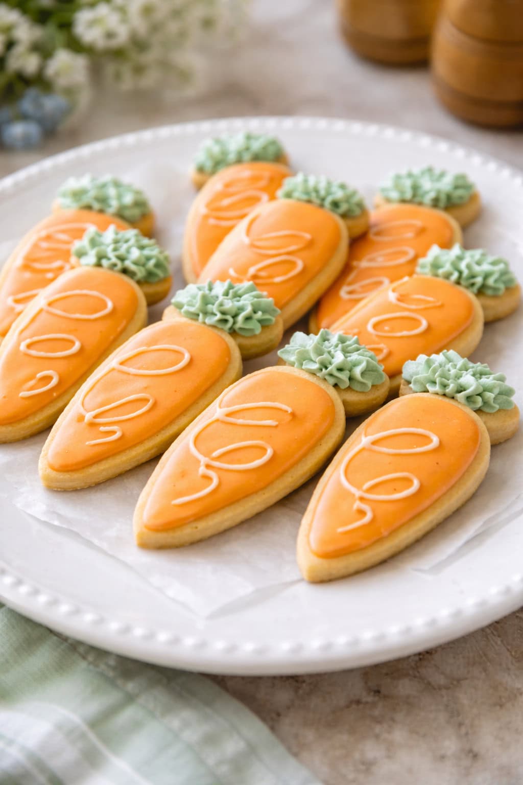 A plate of carrot-shaped sugar cookies, perfect for a bunny baby shower, decorated with orange icing and green frosting tops, arranged neatly on parchment paper.