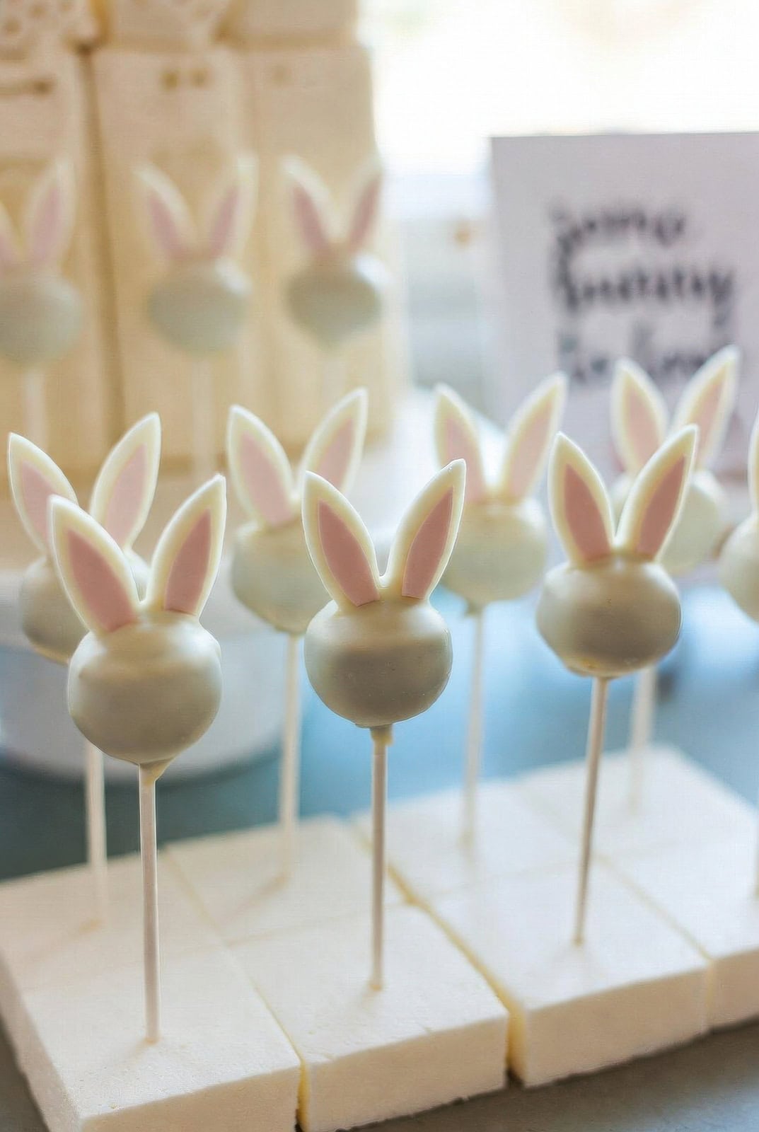 Cake pops decorated as bunny heads with white and pink ears, perfect for a Bunny baby shower, are arranged upright on white foam blocks. In the background, more treats with similar bunny ear decorations add to the festive display.