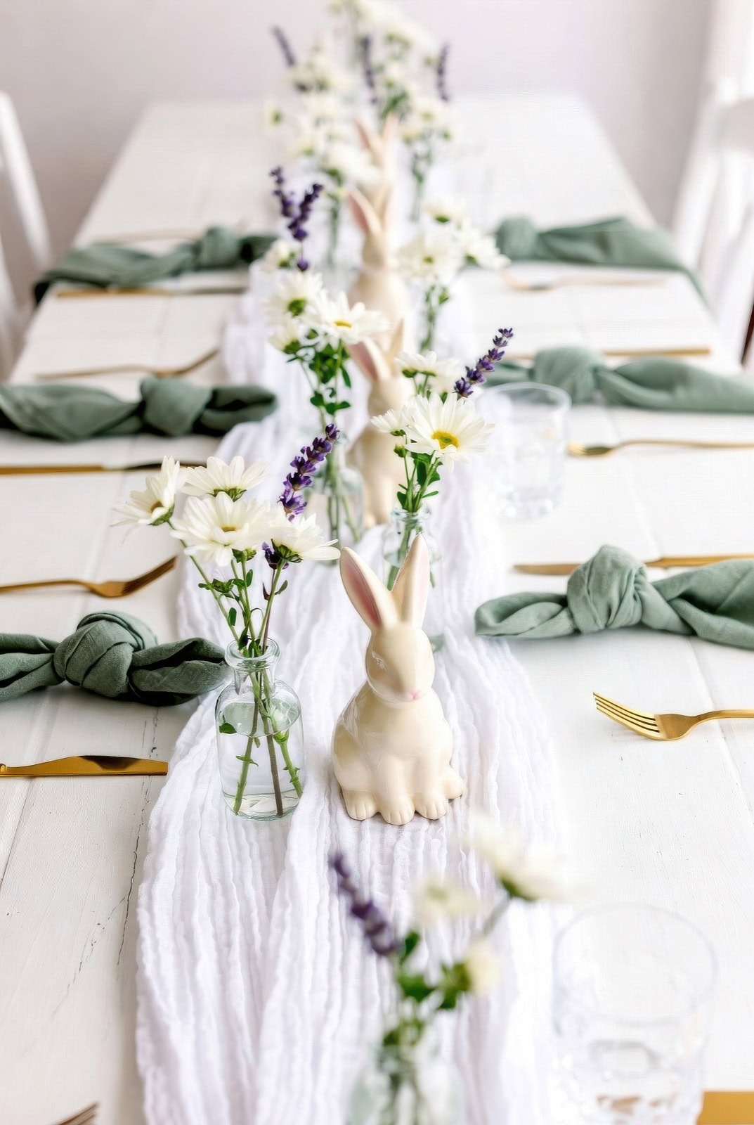 A white table is set for a bunny baby shower with gold cutlery, green napkins, clear glasses, ceramic bunny figurines, and small glass vases holding white daisies and purple flowers along a white fabric runner.