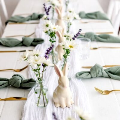 A white table is set with gold cutlery, green napkins tied in knots, small vases of white and purple flowers, and ceramic bunny figurines—perfect for a spring or Bunny baby shower celebration.