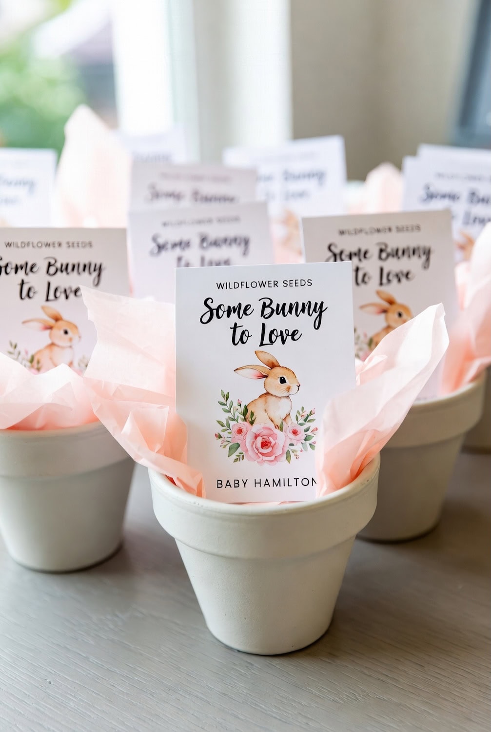 Several white flower pots filled with pink tissue paper, each holding a “Wildflower Seeds: Some Bunny to Love” packet with a bunny and floral design—perfect for a Bunny baby shower. The bottom tag reads “Baby Hamilton.”.