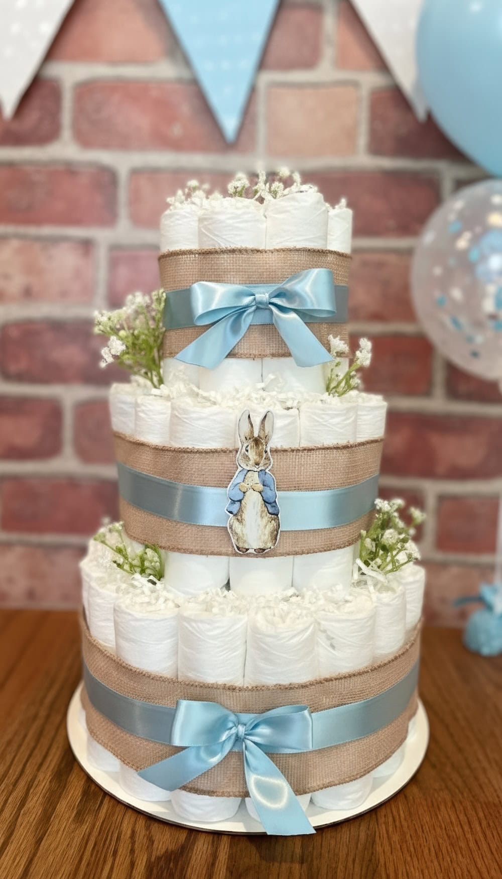 A three-tier diaper cake decorated with blue bows, burlap ribbon, small white flowers, and a Peter Rabbit cutout is displayed on a wooden table—perfect for a bunny baby shower with festive blue and white decorations in the background.