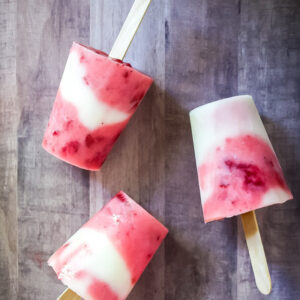 Three homemade strawberry lemonade popsicles, featuring vibrant yogurt and berry swirls in red and white, lie on a wooden surface—two upright and one on its side.