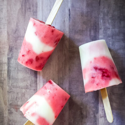 Three homemade strawberry lemonade popsicles, featuring vibrant yogurt and berry swirls in red and white, lie on a wooden surface—two upright and one on its side.