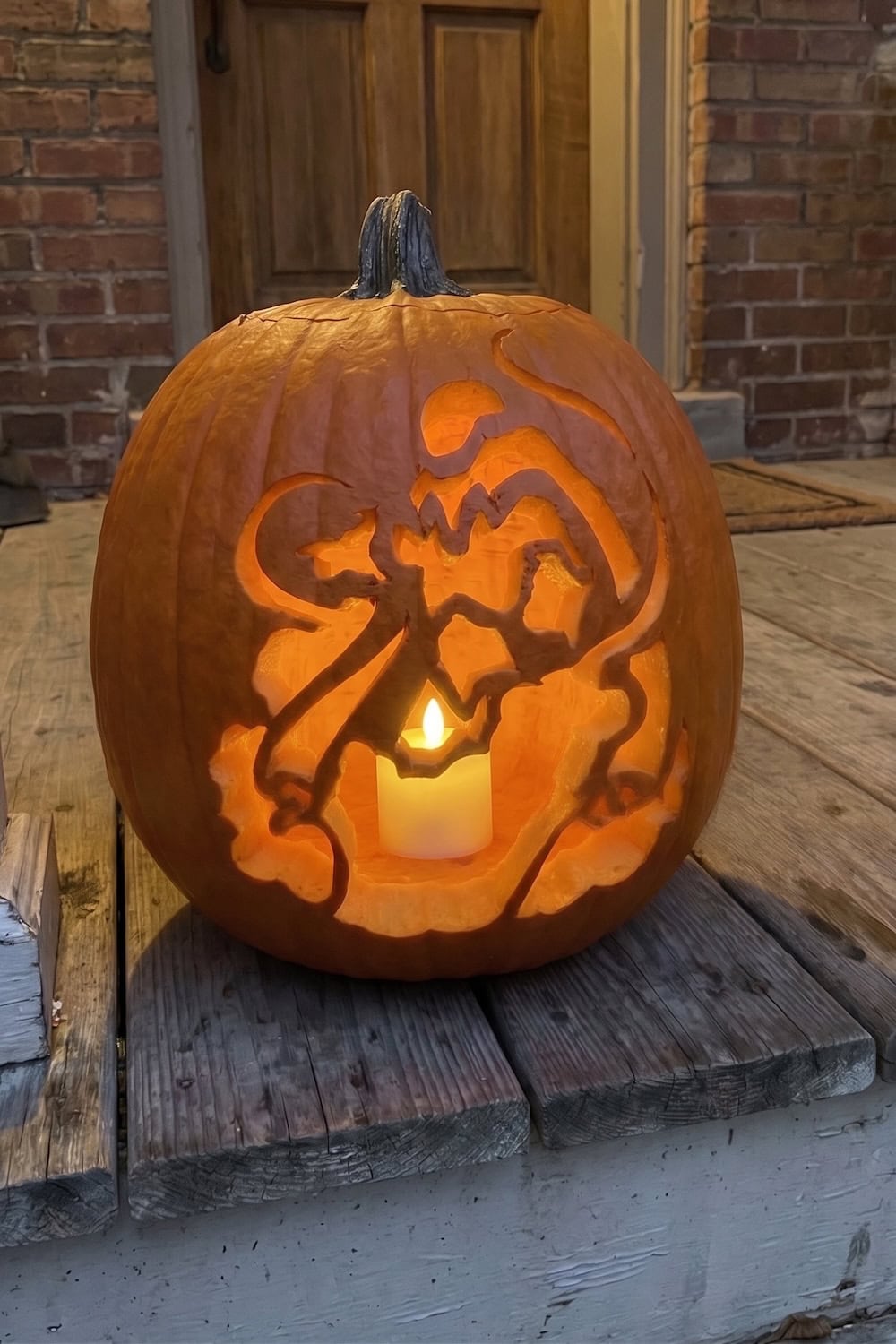 A carved pumpkin featuring a dragon design sits on a wooden porch, illuminated from inside by a lit candle, with a brick wall and door in the background. For those inspired to create their own unique jack-o’-lanterns, consider using Spiderman Pumpkin Stencils and Free Printable Spider-Man Templates for an eye-catching superhero touch this Halloween.