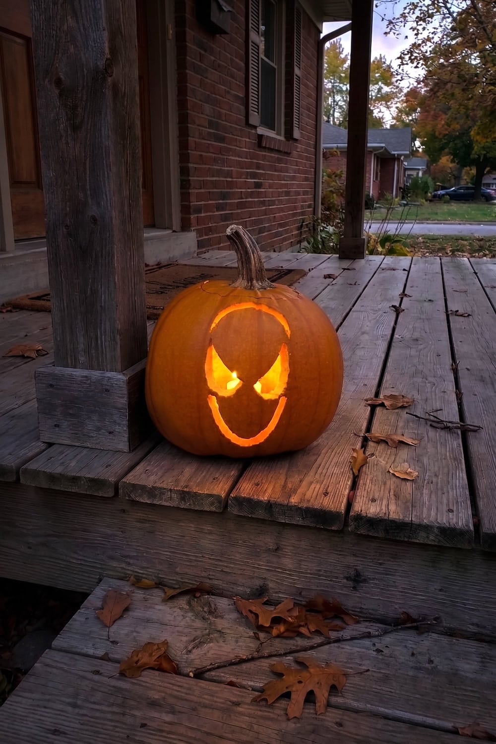 A carved pumpkin with a glowing, sinister face sits on a wooden porch at dusk, surrounded by fallen leaves—its features expertly crafted using Spiderman pumpkin stencils, making it the perfect tribute for fans searching for free printable Spider-Man templates this Halloween.