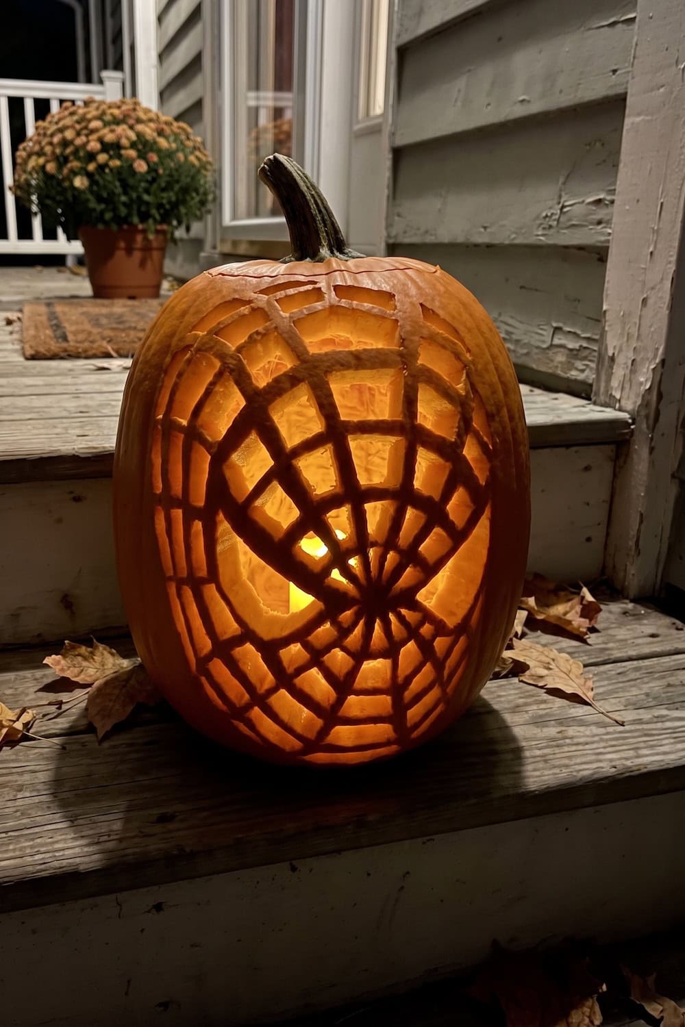A carved pumpkin with a lit candle inside displays a spiderweb design inspired by Spiderman Pumpkin Stencils, sitting on wooden porch steps with fallen leaves and a potted mum in the background.